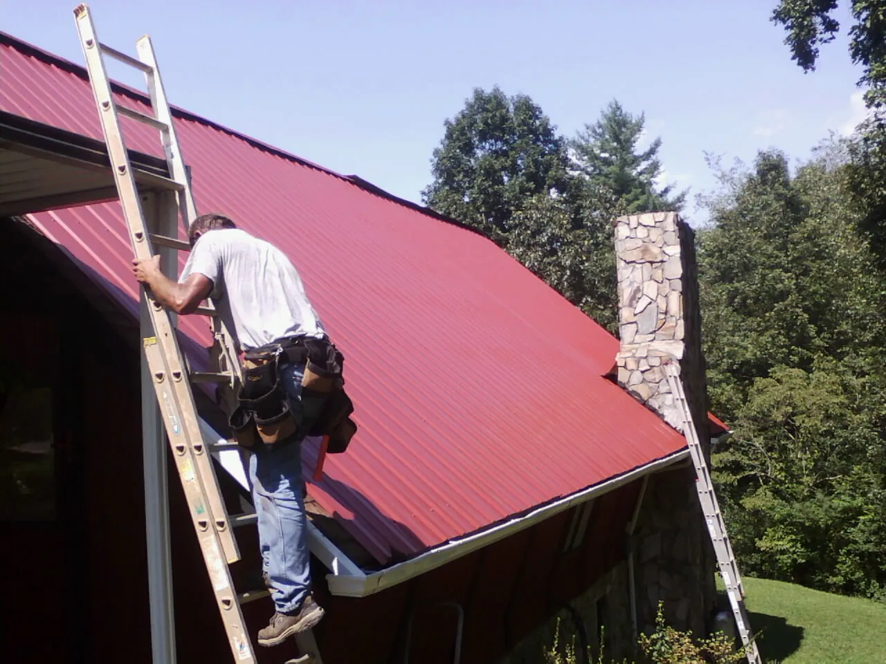 Roofer on a ladder beside a red metal roof and stone chimney — Soffit & Fascia Repair by Roofing Brothers in Dodge Center